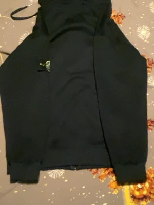 Svart hoodie från Stone Island - Säljer en svart hoodie från Stone Island med den klassiska loggan på ärmen. Hoodien har huva med dragsko och ribbade muddar vid ärmslut och nederkant. Tillverkad i mjukt bomullsmaterial som är skönt att bära.        De L men de är inte L de passar som M/S. Vi kan prata om priset😁