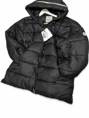 Svart dunjacka med huva från Moncler - ( står S men passar helt klart som en xxl+ för dig som väger mellan 110 och 120kg passar den perfekt)Snygg svart dunjacka från Moncler med huva och dragkedja framtill. Jackan har en quiltad design, två sidofickor med dragkedja och Moncler-logga på ärmen. Perfekt för kalla dagar och riktigt varm tack vare dunfyllningen.