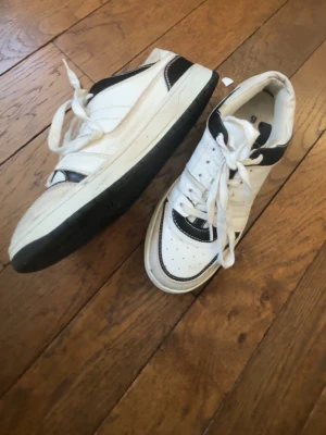 Vita och svarta sneakers med retrostil - Snygga sneakers i vitt och svart med klassisk retrodesign. Skorna har perforerad tå, vita snören och svart detalj runt kragen. 