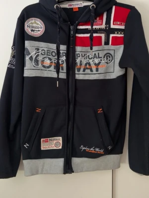  hoodie Geographical Norway - hoodie med dragkedja från Geographical Norway. Har gråa detaljer, broderade loggor, norska flaggan och tryck på både fram- och baksida. Utrustad med huva och två fickor framtill. Snygga orangea detaljer och patchar ger en sportig look.