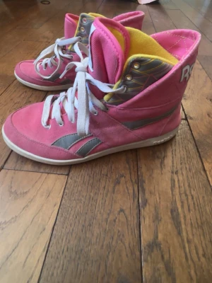 Rosa Reebok high top sneakers - Snygga rosa high top sneakers från Reebok med vita snören och gråa detaljer på sidorna. Insidan är gul och mönstrad, och tungan har Reebok-logga. Skorna har en klassisk retrostil och är tillverkade i mocka och textil.
