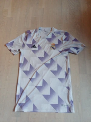 Real Madrid träningströja Adidas XS (Stor I storleken) - Stor i storleken.Snygg Real Madrid träningströja från Adidas i vitt med lila geometriskt mönster. Tröjan har rund hals, korta ärmar och klubbens emblem på bröstet. Perfekt för fotbollsträning eller som supporterplagg.