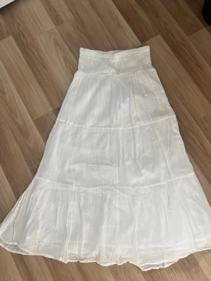 Vit långkjol med smock i midjan - Luftig vit långkjol med smockresår i midjan och flera volanger. Kjolen har en flowy passform och är tillverkad i ett tunt, mjukt tyg som passar perfekt till varma dagar. Enkel och stilren design som är lätt att matcha.
