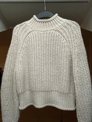 Beige stickad tröja - Snygg stickad tröja från H&M i beige färg med grov struktur och hög ribbad krage. Tröjan har långa ärmar och breda muddar vid ärmslut och nederkant. 