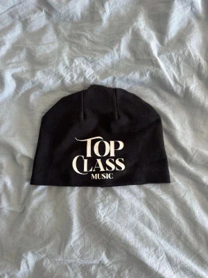 Svart mössa med Top Class Music-tryck - Svart mössa med vit text där det står 'Top Class Music' framtill. Klassisk modell i mjukt tyg, perfekt för att hålla värmen och ge en cool touch till din outfit.