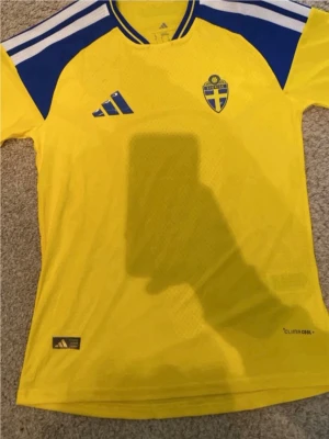 Svensk landslagströja Adidas gul/blå - Gul och blå fotbollströja från Adidas med Sveriges landslagsemblem på bröstet. Tröjan har ClimaCool-material som andas, korta ärmar och klassiska Adidas-ränder på axlarna. Perfekt för match eller träning.