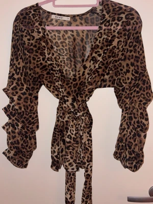 Leopardmönstrad omlottblus från NA-KD - Trendig omlottblus från NA-KD i transparent, lätt material med leopardmönster. Blusen har långa volangärmar och knyts i midjan för en snygg siluett. Perfekt för dig som vill sticka ut med ett djurmönstrat plagg.