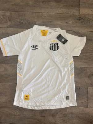 Vit Santos FC matchtröja från Umbro - Vit fotbollströja från Umbro med Santos FC:s klubbmärke på bröstet. Tröjan har svarta och gula detaljer, korta ärmar och V-ringning. Tillverkad i lätt och ventilerande material, perfekt för träning eller match.