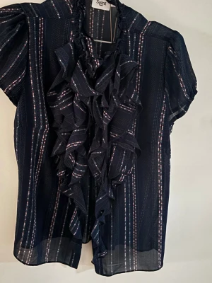Mörkblå randig blus med volanger - Snygg mörkblå blus från Saint Tropez med tunna vertikala ränder i vitt och rött. Blusen har korta puffärmar och volangdetaljer framtill som ger en cool och trendig look. Tillverkad i ett lätt och transparent material som känns luftigt. Helt ny med prislapp på där den kostar 449 kr.💕säljer för att de var fel storlek men super fin.