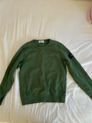 Grön tröja från Stone Island Junior  - Snygg grön tröja från Stone Island junior. Den har använts ganska mycket men den är i ett väldigt fint skick. Den har ett litet hål på ena armen (se sista bilden) men det går att laga enkelt och jag kan även laga det fint innan jag skickar tröjan. Tröjan är i storlek 10 år (142). Priset kan diskuteras och noggrannare bilder kan skickas