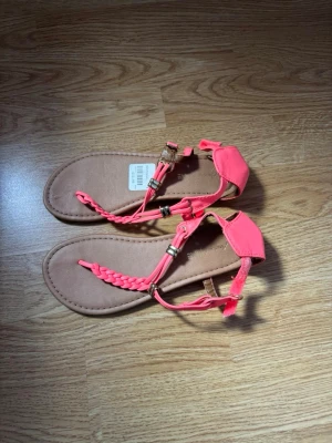 Korallfärgade flätade sandaler - Snygga sandaler i korallrosa med flätad rem och guldfärgade detaljer. De har en platt sula i beige och justerbart spänne vid ankeln. 