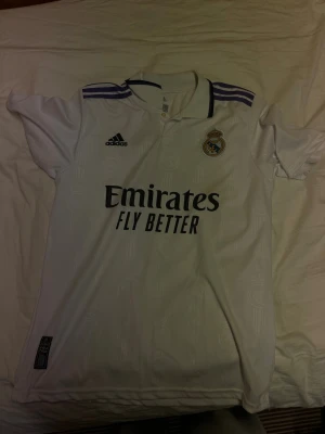 Real Madrid matchtröja Benzema #9 - Vit Real Madrid matchtröja från Adidas med lila detaljer på axlarna. Tryck med klubbmärke, Adidas-logga och sponsor på framsidan. På ryggen står det 'Benzema' och nummer 9. Perfekt för fotbollsfans!