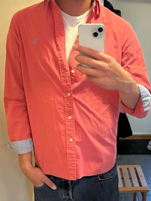 Korallröd skjorta från Ralph Lauren - Snygg korallröd skjorta från Ralph Lauren i 100% bomull med stretch. Skjortan har klassisk krage, vita knappar och långa ärmar med ljusblå insida på manschetterna. Perfekt för en stilren och avslappnad look.