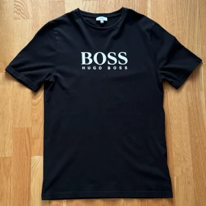 Hugo Boss T-Shirt - Klassisk Hugo Boss T-shirt i svart med vit logga framtill. Storlek 16M / ungdom, motsvarar ungefär herr XS–S beroende på önskad passform. Stilren och lättmatchad till vardags.