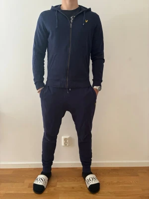 Marinblå loungewear-set från Lyle & Scott - Marinblå träningsdress från Lyle & Scott med zip-hoodie och matchande byxor. Hoodien har dragkedja, fickor och logga på bröstet. Byxorna har resår i midjan och muddar vid benslut. Perfekt för gymmet eller chill hemma. Sitter som storlek M