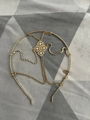 Guldigt diadem med kedjedetaljer - Snyggt diadem i guldfärgad metall med tunna kedjor och en dekorativ, utskuren detalj i mitten. Diademet har flera kedjor som hänger ner och ger en bohemisk och unik look. Perfekt accessoar för att lyfta din outfit.