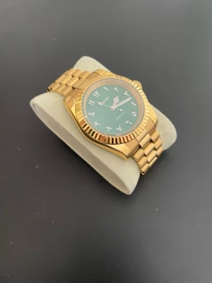 Seiko guldig klocka med grön urtavla - Stilren Seiko klocka i guldigt metallarmband och boett. Urtavlan är grön med arabiska siffror och har en snygg textur. Automatisk rörelse och klassisk design som sticker ut med sin färgkombination. Obs: Fungerar ej!!