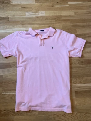 Ljusrosa pikétröja från Gant - Snygg ljusrosa pikétröja från Gant med klassisk krage och korta ärmar. Tröjan har broderad logotyp på bröstet och är tillverkad i mjuk bomull. Perfekt för en avslappnad och stilren look. Skick 9/10