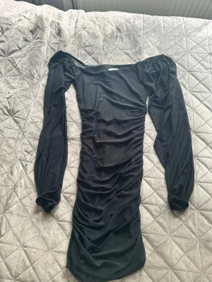 Svart långärmad klänning från Nelly - Svart figurnära klänning från Nelly med långa puffärmar och rynkade detaljer längs hela klänningen. Offshoulder-modell som ger en trendig look. Perfekt för dig som gillar stilrena och snygga outfits.