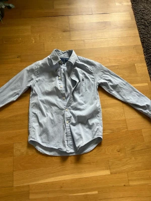 Ljusblå skjorta från Ralph Lauren - Klassisk ljusblå skjorta från Ralph Lauren med broderad logga på bröstet. Skjortan har lång ärm, knappar framtill och en tidlös passform. Perfekt för en stilren look.