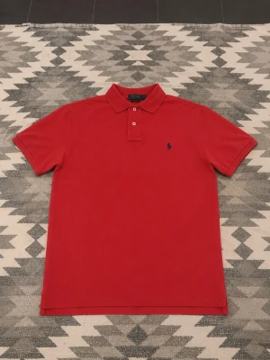Röd pikétröja från Ralph Lauren - Klassisk röd pikétröja från Ralph Lauren med korta ärmar och krage. Tröjan har två knappar vid halsen och den ikoniska broderade loggan på bröstet. Perfekt för en stilren och avslappnad look.