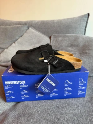 Svarta Birkenstock Boston tofflor - Säljer ett par svarta Birkenstock Boston tofflor i mocka med justerbart spänne och klassisk korksula. Perfekta för dig som gillar stilrena och bekväma skor. Modellen är tidlös och passar till många olika outfits.