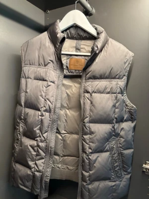 Grå dunväst från Massimo Dutti - Snygg grå dunväst från Massimo Dutti med hög krage och quiltad design. Västen har dragkedja framtill och två fickor på sidorna. Perfekt för lager-på-lager och ger en stilren look. Broderad logga i nacken.
