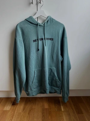 Grön hoodie från Better Bodies - Grön hoodie med svart Better Bodies-tryck på bröstet. Hoodien har en stor magficka, huva med dragsko och långa ärmar. Materialet är mjukt och bekvämt, perfekt för att chilla eller träna i.