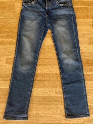 Blå raka jeans med slitningar - Säljer ett par klassiska blå jeans med snygga slitningar framtill. Jeansen har fem fickor och normalhög midja. Perfekta för en avslappnad och trendig look. Materialet är denim i bomull med lite stretch för extra komfort. 