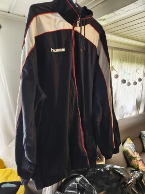 Svart träningsjacka från Hummel/xxl - Snygg svart träningsjacka från Hummel med vita och röda detaljer. Jackan har dragkedja framtill, hög krage och klassiska Hummel-loggor på ärmarna. Tillverkad i lätt och smidigt syntetmaterial, perfekt för sport och träning.