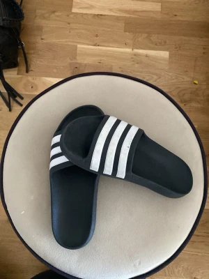 Svarta Adidas badtofflor med vita ränder - Klassiska svarta badtofflor från Adidas med tre vita ränder över foten. Tillverkade i syntetmaterial med en enkel slip-on design. Perfekta för stranden eller gymmet och har en sportig look som aldrig går ur tiden.
