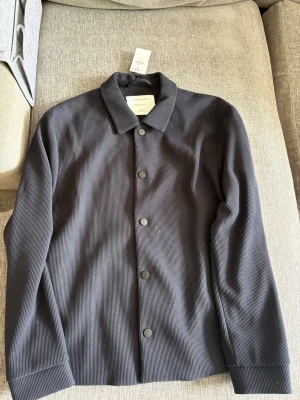 Svart overshirt från Primark Edit - Stilren svart overshirt från Primark Edit i regular fit. Jackan har en klassisk krage, knäppning framtill med svarta knappar och är tillverkad i ett ribbat tyg som ger en modern känsla. Perfekt att slänga över en t-shirt för en avslappnad look.