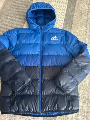 Blå pufferjacka från Adidas 13-14 Y/ 164 cm - Snygg blå och mörkblå pufferjacka från Adidas med huva och dragkedja framtill. Jackan har två sidofickor med dragkedja och Adidas-logga på bröstet. Tillverkad i glansigt syntetmaterial, perfekt för kalla vinterdagar.