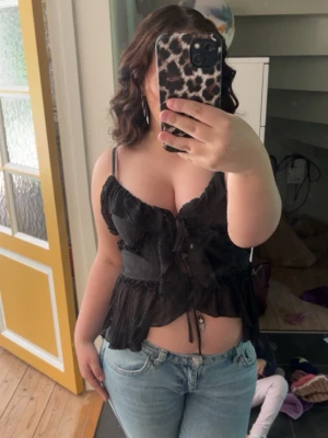 Svart linnetopp med volang och knyt - Snygg svart linnetopp med tunna axelband, volangdetaljer och knyt framtill. Toppen har en lätt plisserad look och är croppad, perfekt till jeans eller kjol. Materialet känns lätt och luftigt.