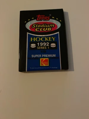 Topps Stadium Club Hockeykort 1992 - Ett oöppnat paket med Topps Stadium Club hockeykort från 1992, Series 1. Förpackningen är svart och blå med tydliga loggor och text, samt Kodak Imaging Technology-märke. Perfekt för samlare av sportkort och nostalgi från 90-talet.