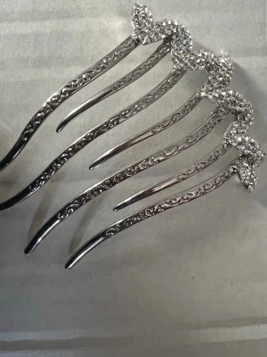 Silverfärgad hårkam med strasshjärtan - Snygg hårkam i silverfärgad metall med sex tänder och dekorativa hjärtan upptill, täckta av glittrande strasstenar. Kammen har graverade detaljer längs tänderna som ger en lyxig känsla. Perfekt accessoar för att lyfta din frisyr.