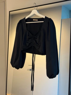 Svart croppad topp med puffärmar - Svart croppad topp med fyrkantig ringning med långa puffärmar och  rynkdetalj framtill med knytband. 