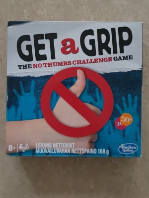 Brädspel - Get a Grip är ett roligt sällskapsspel där du ska klara utmaningar utan att använda tummarna. Spelet kommer i en fyrkantig kartong med blå, röd och vit design och innehåller bland annat modellera, kort och tumhandskar. Perfekt för spelkvällar med vänner eller familj.