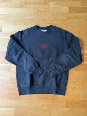Svart sweatshirt från Han Kjøbenhavn - Svart sweatshirt från Han Kjøbenhavn med rund hals och röd broderad logga på bröstet. Klassisk passform och ribbade muddar vid ärmslut och nederkant. Perfekt för en stilren och avslappnad look.