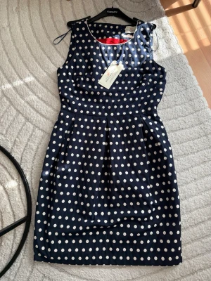 Marinblå prickig klänning Holly & White - Snygg marinblå klänning från Holly & White med vita prickar och ärmlös design. Klänningen har en klassisk, figurnära passform och markerad midja. Perfekt för dig som gillar retrovibbar och tidlös stil.