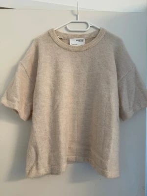 Beige stickad t-shirt från Selected femme - Säljer en beige stickad t-shirt från Selected femme med rund halsringning och korta ärmar. Tröjan har en avslappnad passform och är gjord i mjukt stickat material, perfekt för lager på lager eller sommarens svalare dagar.