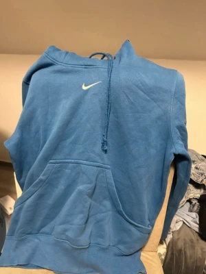 Blå Nike hoodie med logga - Blå hoodie från Nike med vit broderad Swoosh-logga på bröstet. Klassisk modell med känguruficka och justerbar huva med dragsko. Mjuk bomullsblandning som är skön att ha på sig. Perfekt för en avslappnad streetstyle-look.