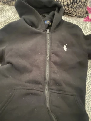 Svart hoodie med dragkedja från Polo - Svart hoodie med dragkedja från Polo Ralph Lauren. Klassisk design med vit broderad logga på bröstet och kängurufickor framtill. Mjuk och skön bomullsblandning, perfekt för en avslappnad stil.
