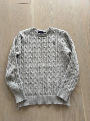 Polo Ralph lauren grå knitwear - Kabelstickad tröja i ljusgrått från Polo Ralph Lauren med rund halsringning och ribbade muddar. Klassisk broderad logga på bröstet. Tillverkad i mjukt material som passar perfekt för lager på lager och ger en stilren look.