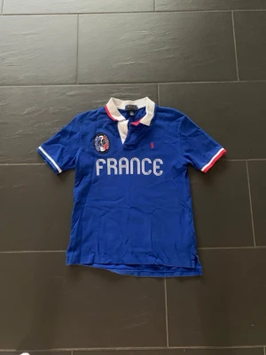 Blå pikétröja France Polo Ralph Lauren - Blå pikétröja från Polo Ralph Lauren med vit krage och FRANCE-tryck på bröstet. Broderad logga och emblem på vänster sida. Röd och vit detalj på ärmslut och krage. Perfekt för dig som gillar grisch stil.