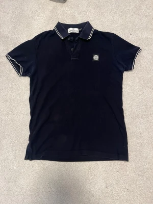 Svart polotopp från Stone Island - Säljer en svart polotopp från Stone Island med klassisk krage och knappslå. Toppens krage och ärmslut har vita kontrastlinjer och logotypen är broderad på bröstet. Perfekt för dig som gillar stilrena och sportiga plagg.