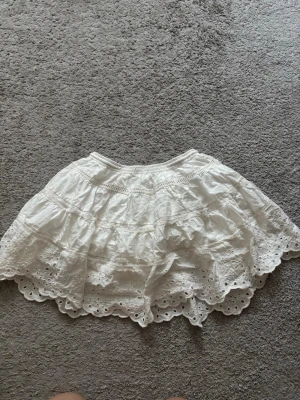 Vit volangkjol med broderi från Zara - Supersöt vit kortkjol från Zara med volanger och broderade detaljer. Kjolen har elastisk midja och vackert hålbroderi längs nederkanten som ger en romantisk känsla. Perfekt för varma dagar och lätt att matcha med olika toppar.