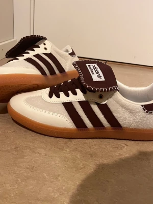 Adidas Wales Bonner Samba sneakers - Snygga Adidas Samba sneakers i samarbete med Wales Bonner. Skorna är vita med mörkbruna detaljer, klassiska tre ränder och brun sula i gummi. Ovandelen är i läder och mocka med unik tunga och kontrastsömmar. Perfekta för dig som gillar stilrena och trendiga sneakers.