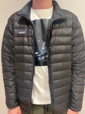 Svart dunjacka från Patagonia - Snygg svart dunjacka från Patagonia med quiltad design och dragkedja framtill. Jackan har en klassisk passform och är lätt vadderad, perfekt för kyliga dagar. Diskret logga på bröstet och hög krage för extra värme. OBS!! Dragkedjan behöver fixas och den har små hål vid ärmen. Hör av er gällande bilder eller funderingar!!