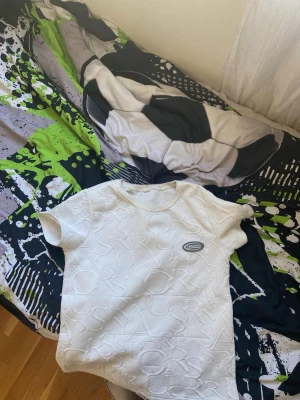 Vit t-shirt från Sportswear - Vit t-shirt med diskret präglat mönster av bokstäver över hela tyget och rund hals. Liten logotyp-patch på bröstet. Klassisk passform och korta ärmar, perfekt för en clean och stilren look.